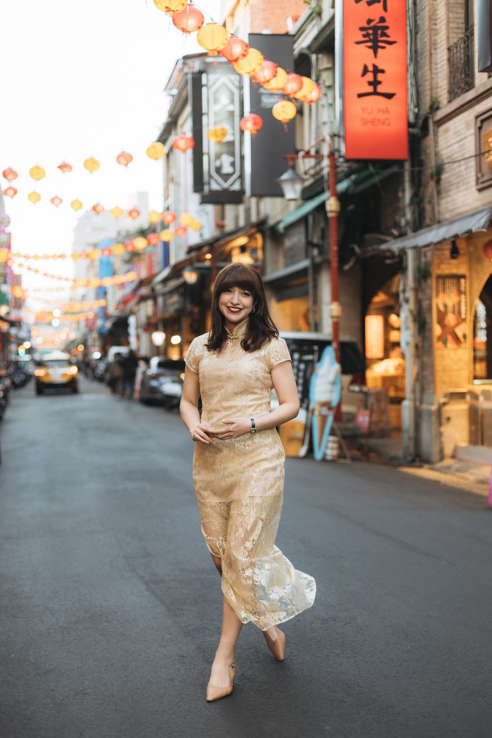 Woman in gold qipao dress walking on a lantern-lined street in an Asian city at dusk.