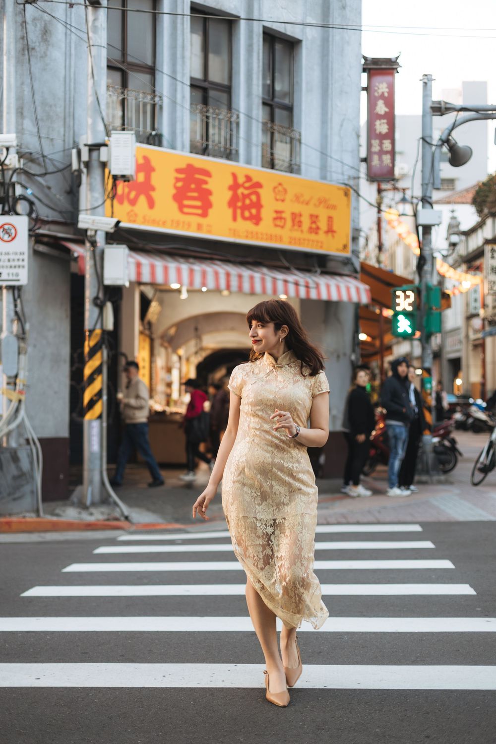 Woman in elegant floral qipao dress crossing a bustling Asian street market at dusk.