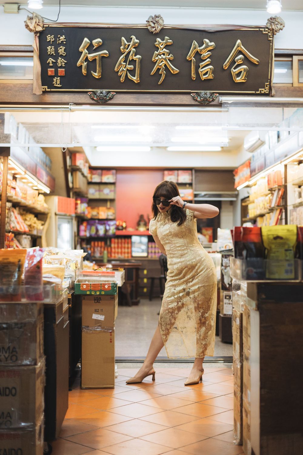 Woman in gold qipao dress photographing inside a traditional Chinese herbal medicine shop with ornate signage.