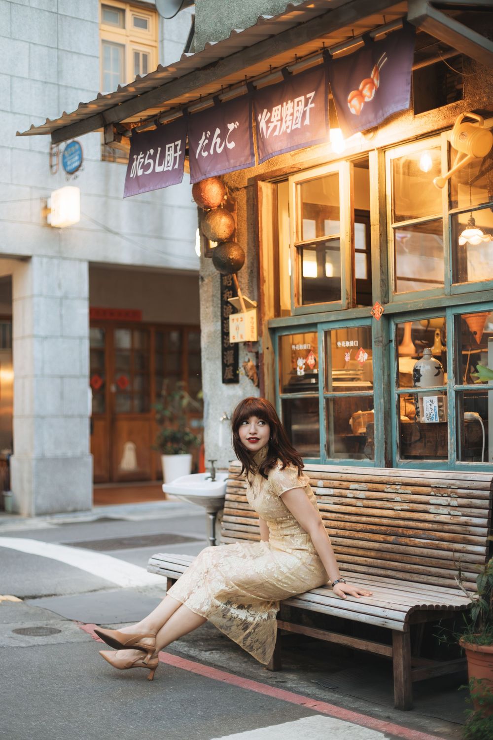 Woman in floral dress sitting on wooden bench outside a vintage Japanese restaurant with lanterns and noren curtains.