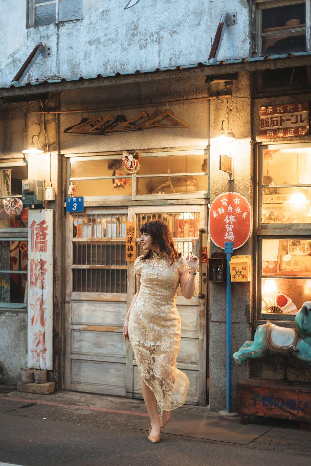 Woman in lace dress poses outside a vintage Asian shop with glowing signs at dusk.