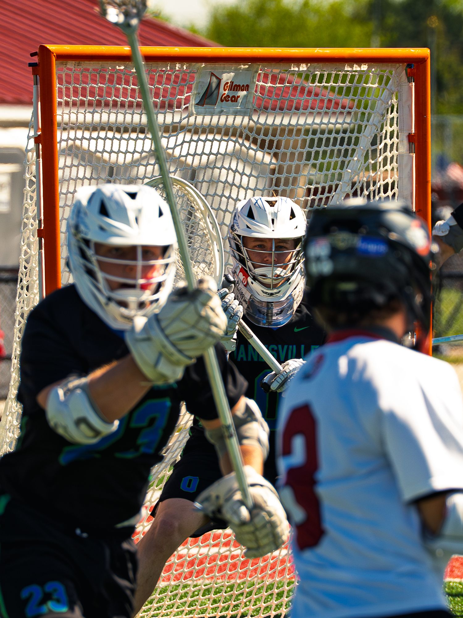 Janesville lacrosse goalie set in the crease behind a defender as an opposing attacker circles the net in daytime sun.
