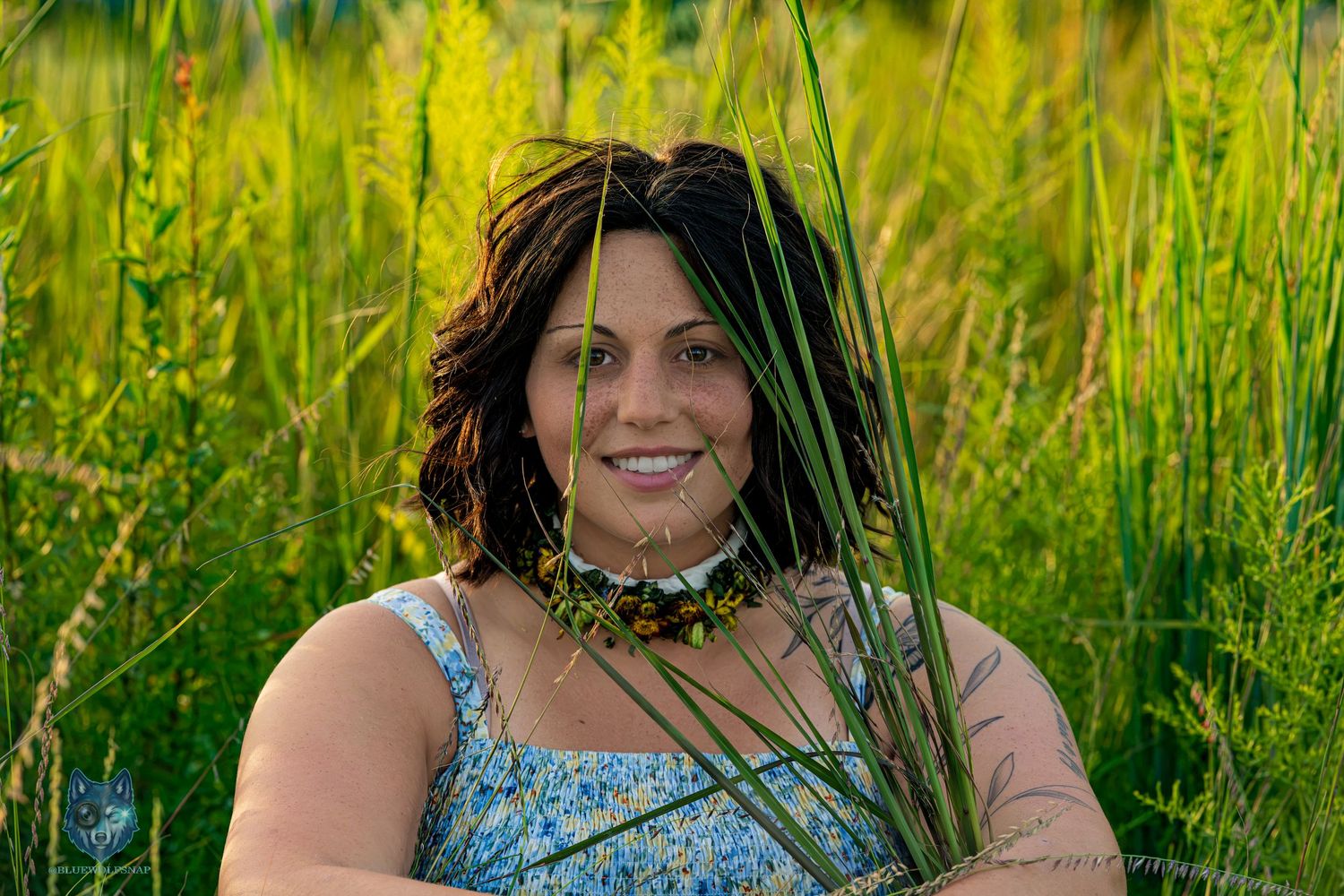 Smiling woman in blue floral dress sits surrounded by lush green tall grass. Chi.Shooters meet gabbsbosh
