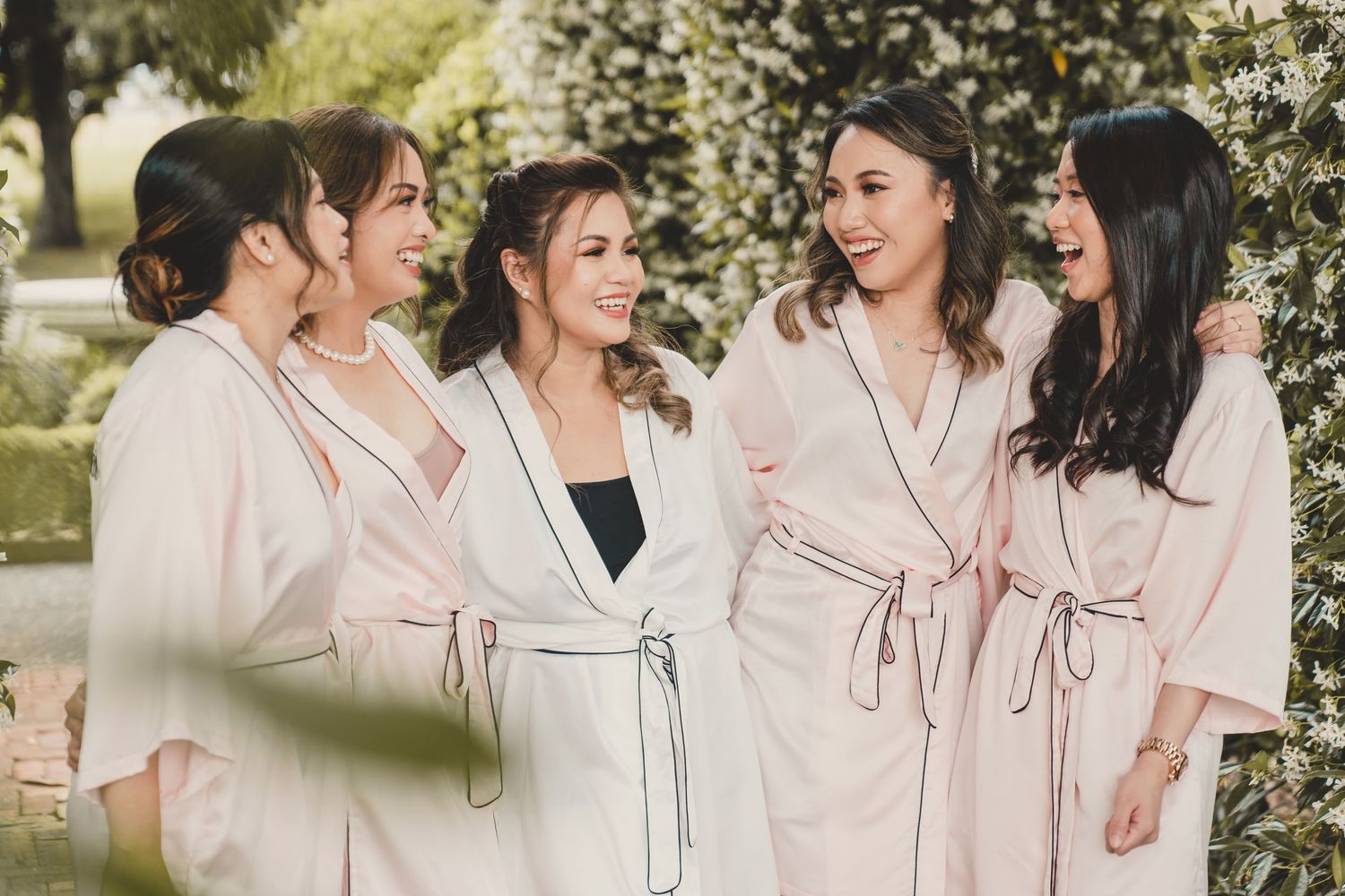 Bride and bridesmaids laughing together in matching blush pink robes outdoors among lush greenery.