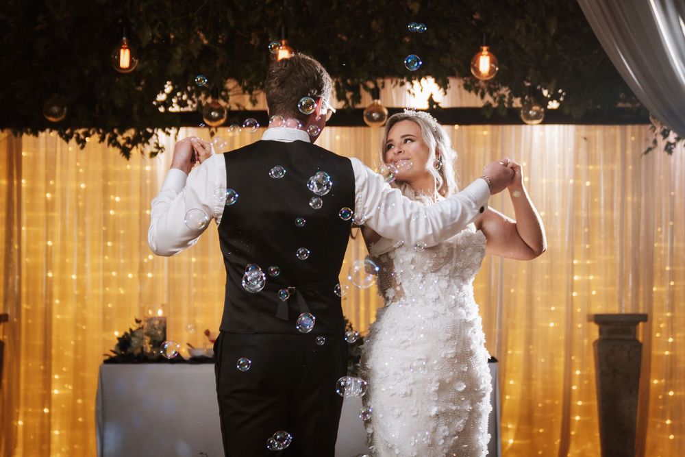 Bride and groom sharing their first dance surrounded by bubbles in front of a warm fairy light backdrop.