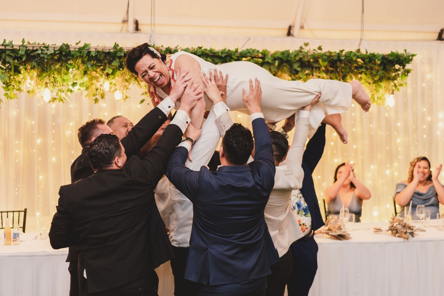 Groomsmen lifting the groom in the air during a joyful wedding reception with greenery and fairy lights.