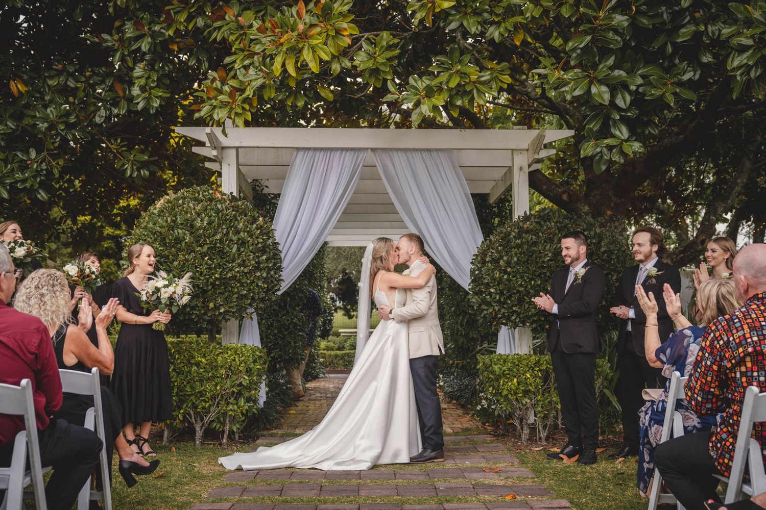Bride and groom share first kiss at outdoor garden wedding ceremony with white draped arbor and guests applauding.