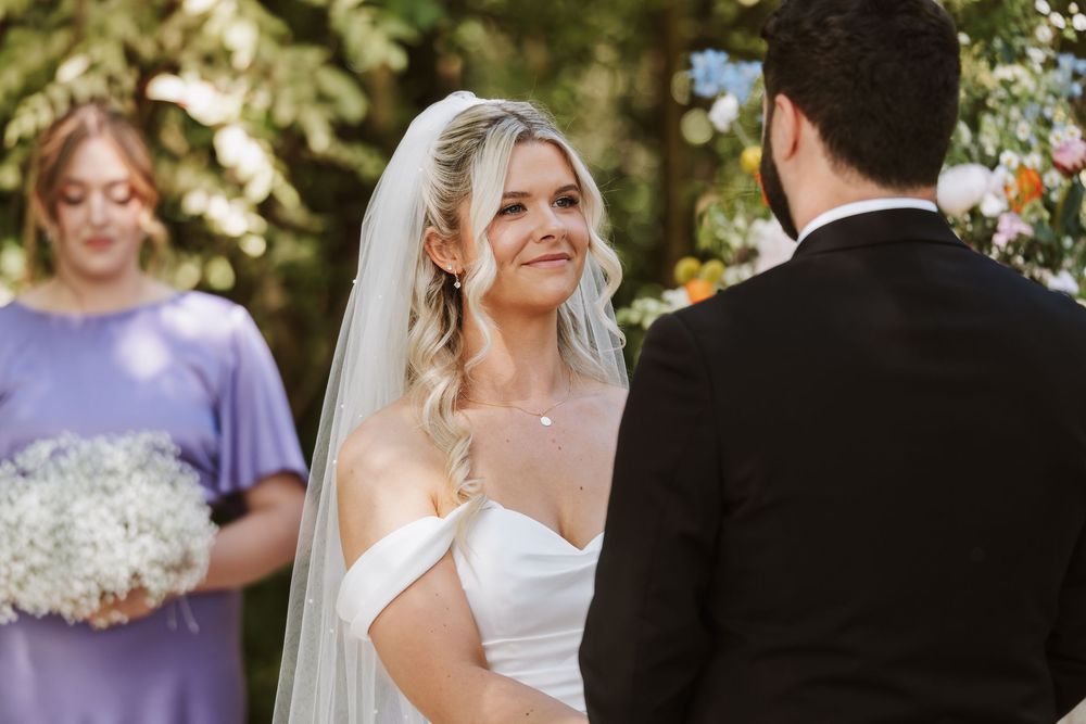 Bride in off-shoulder gown and veil smiles at groom during outdoor wedding ceremony with bridesmaid in purple.