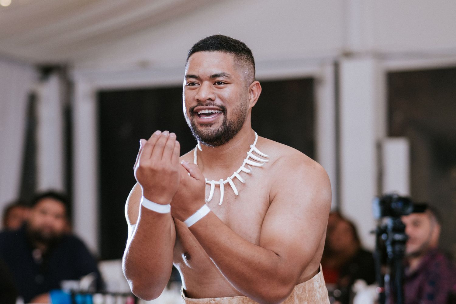 A Polynesian man in traditional attire claps and smiles during a cultural performance at an indoor event.