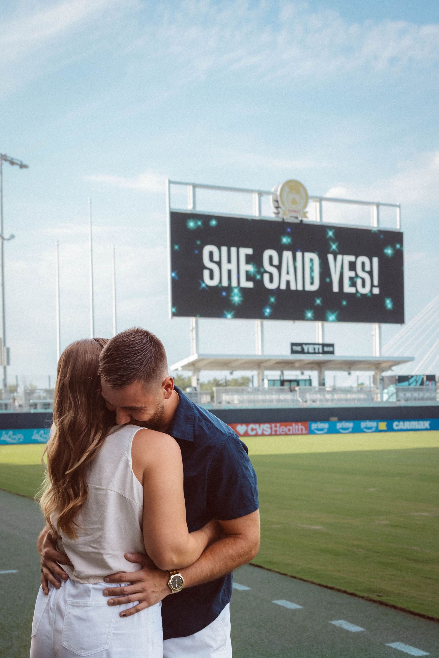 A man and woman embrace in front of a jumbotron reading "She said yes!" during their surprise proposal at CPKC stadium in Kansas City.