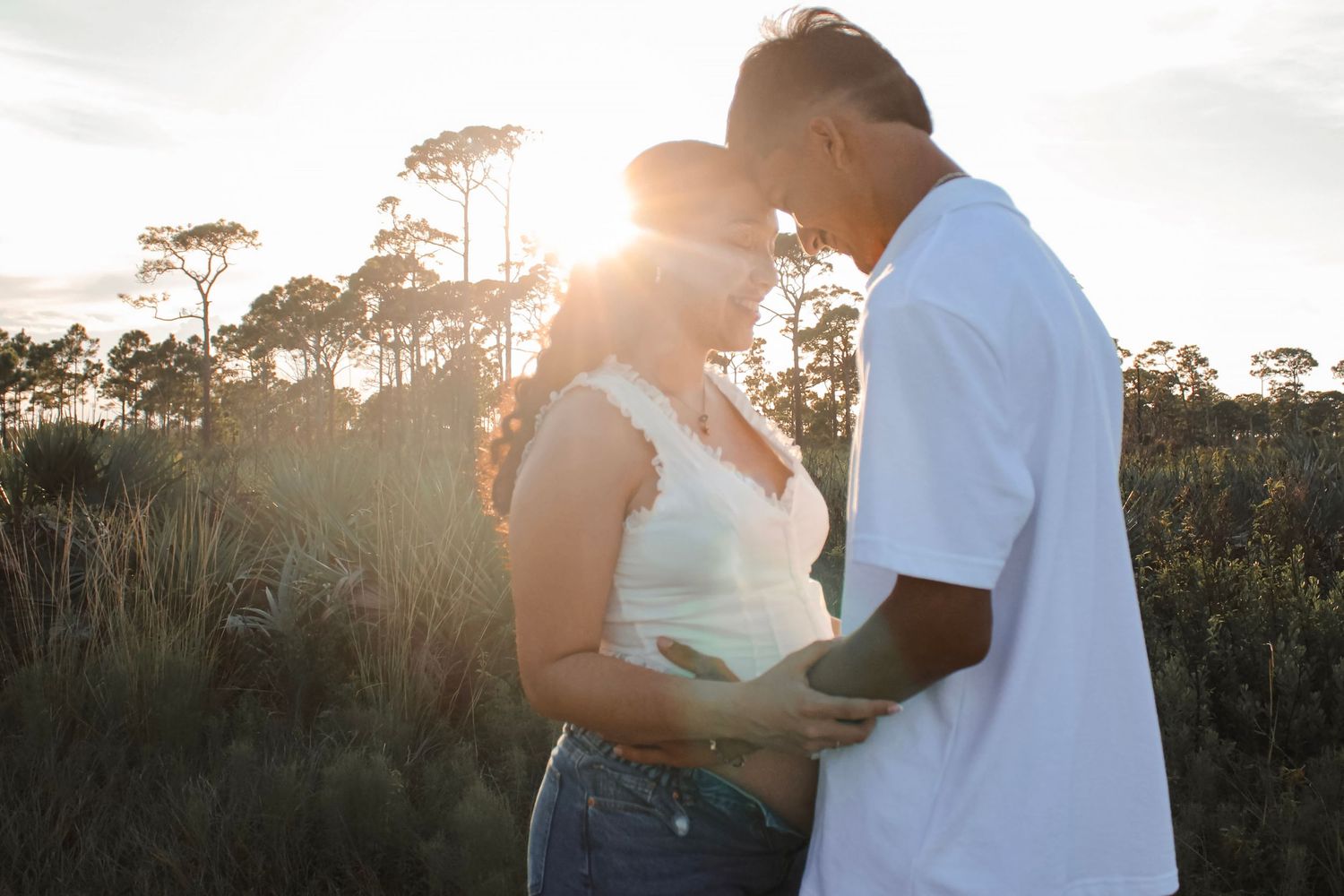Couple embracing at sunset in tall grass field during golden hour outdoor maternity photoshoot.
