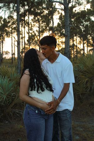 Pregnant couple kissing romantically in pine forest during golden hour sunset.