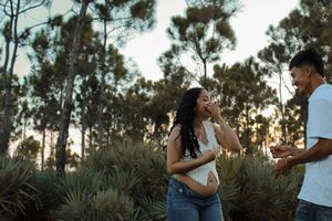 Woman covers mouth in surprise as man shows engagement ring outdoors in pine forest.