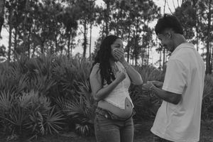 Black and white photo of man proposing to emotional woman in pine forest setting.