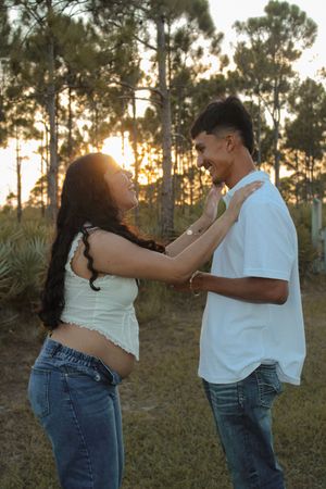 Happy pregnant couple smiling and embracing in golden sunset pine forest.