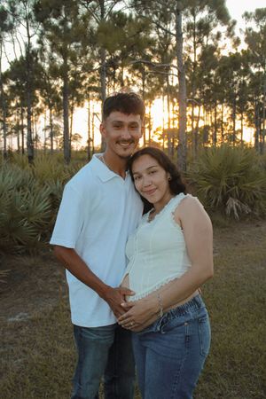 Couple posing outdoors at sunset in white shirts and jeans, woman showing baby bump among pine trees.
