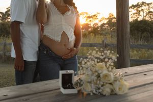 Pregnant couple at sunset with engagement ring box and white flower bouquet on wooden table.