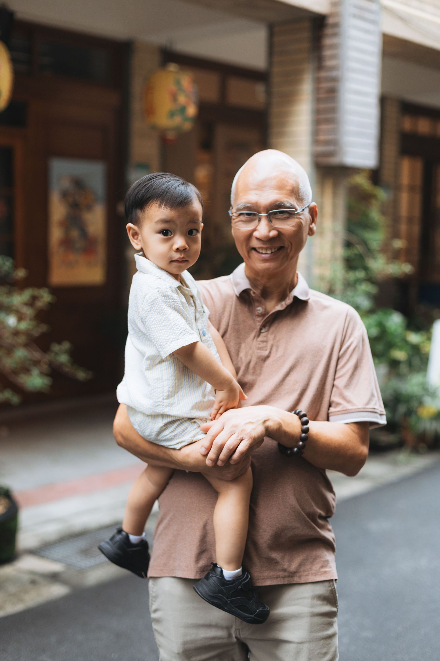 Smiling elderly Asian man holding a young toddler boy outdoors in an urban alley setting.