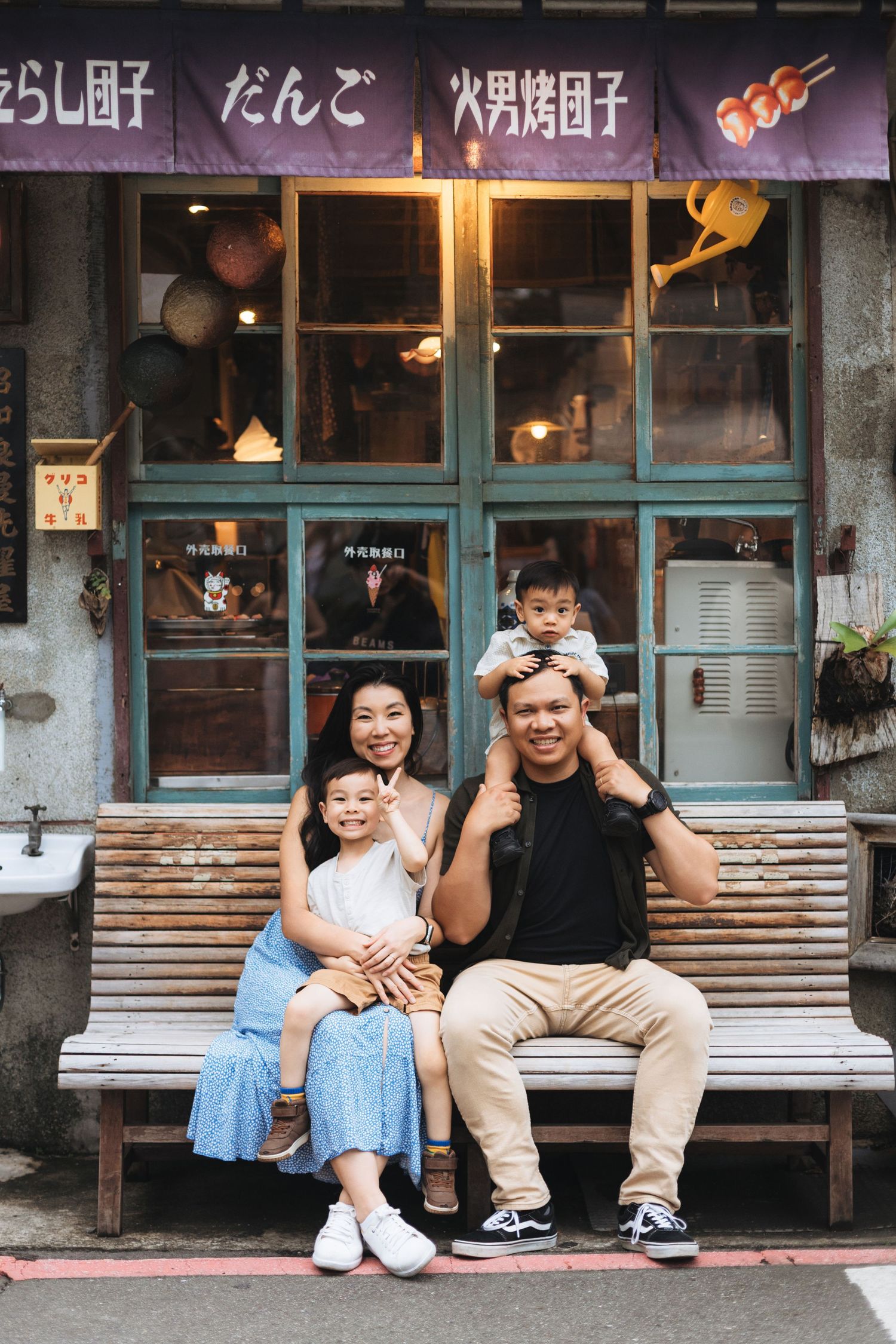 Asian family of four posing on bench outside traditional Japanese dango shop, parents with two young children smiling.