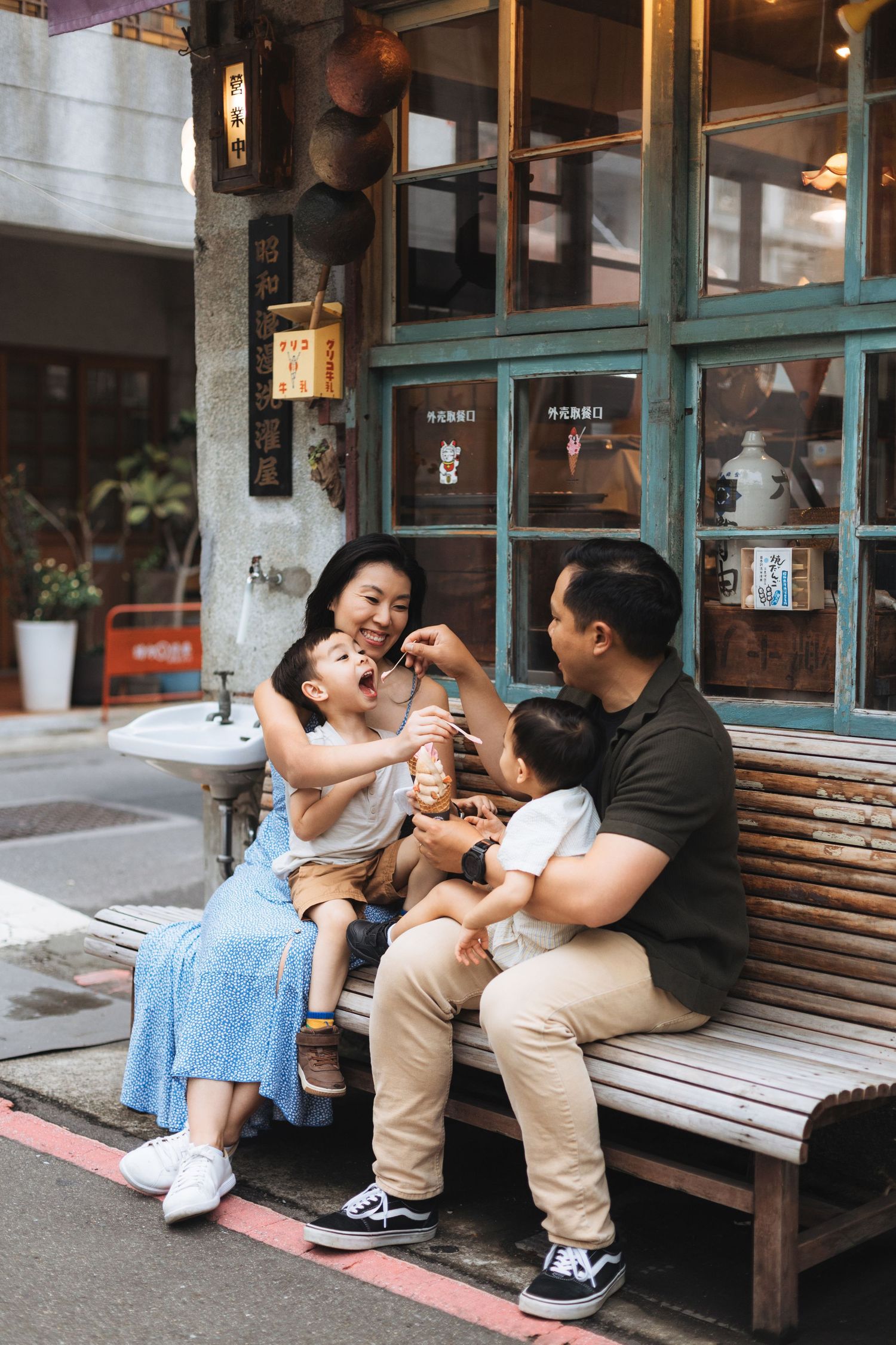 Asian family of four sharing ice cream on a bench outside a vintage restaurant, parents and two young children smiling.