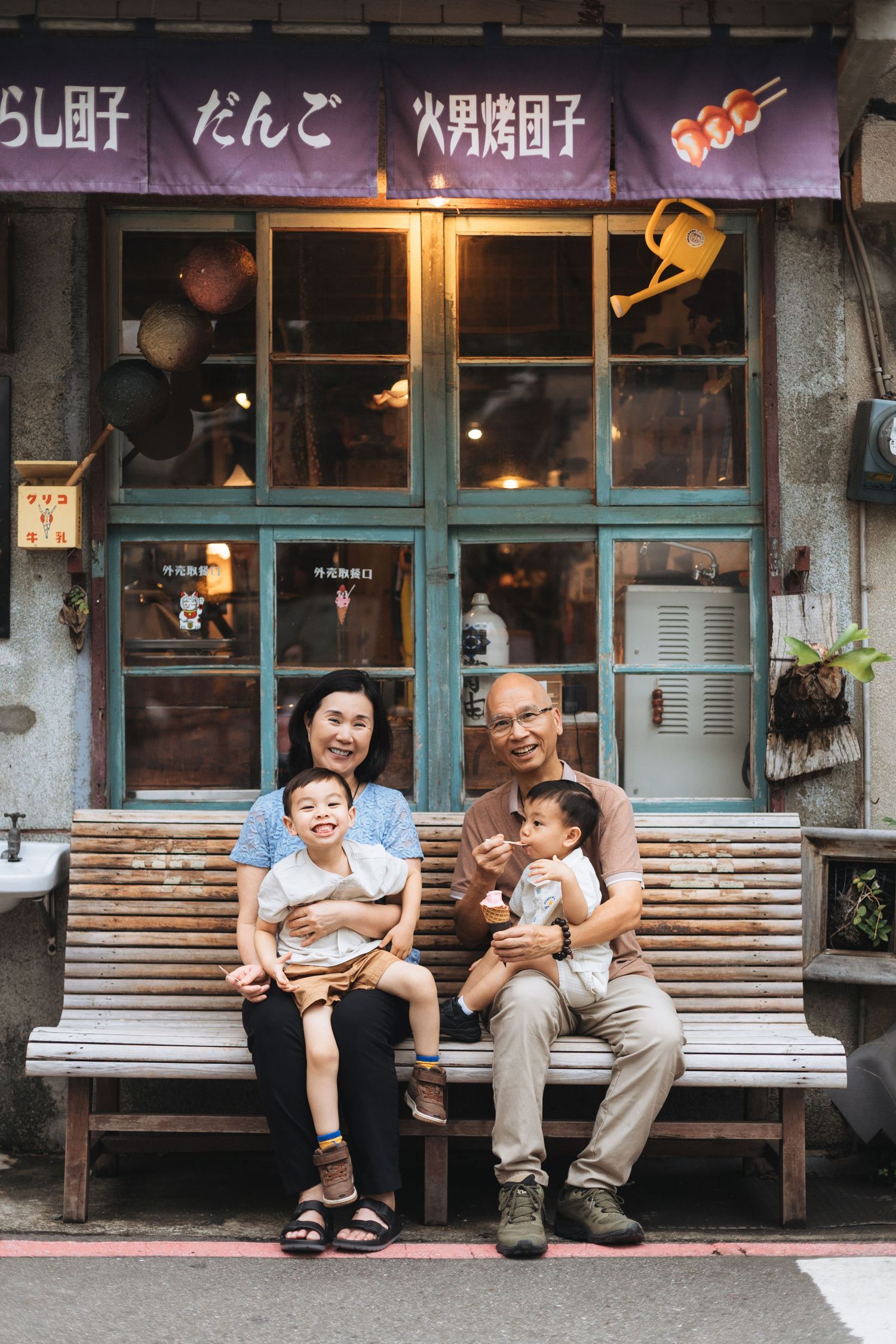 Grandparents sitting on bench outside Japanese dango shop with two young grandchildren, smiling warmly.