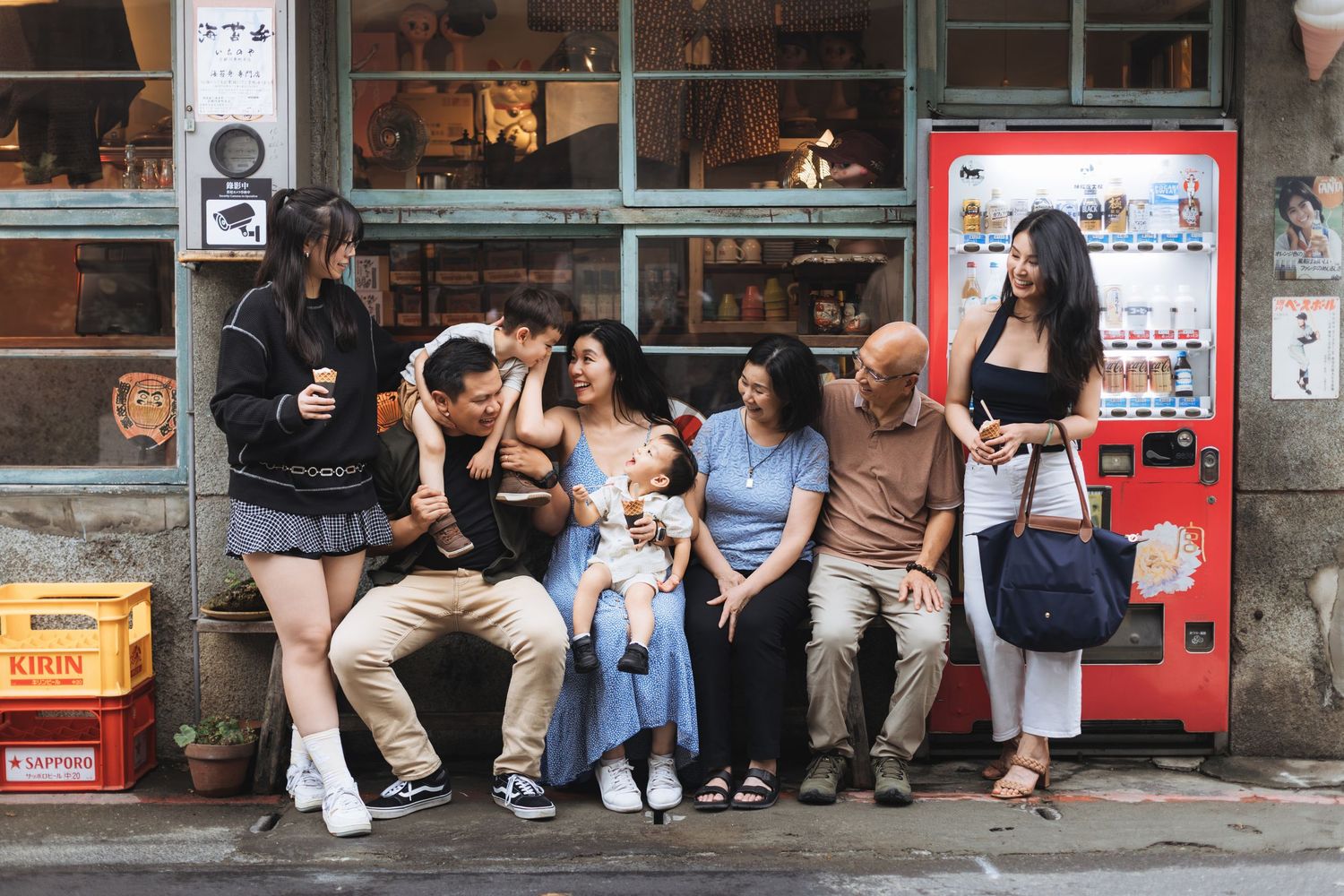 Multi-generational Asian family sitting together outside a vintage shop near a red vending machine, sharing a joyful moment.