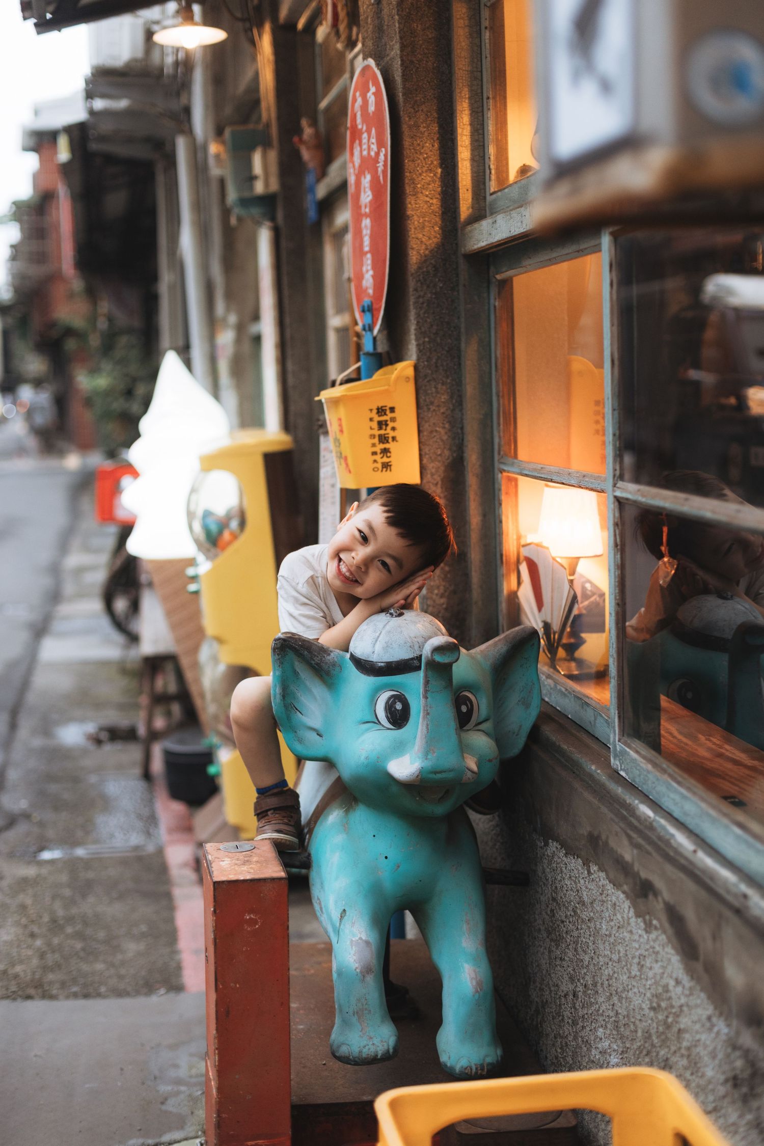 Child leaning on a teal elephant ride-on toy outside a charming Asian street shop with warm lighting.