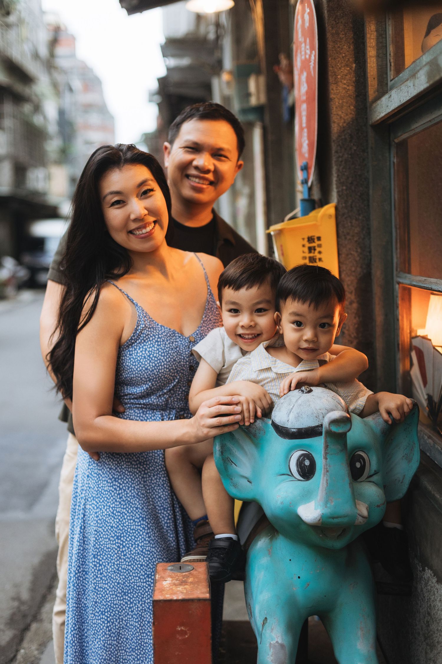 Happy Asian family of four posing on a charming urban alley, two young boys sitting on a teal elephant ride.
