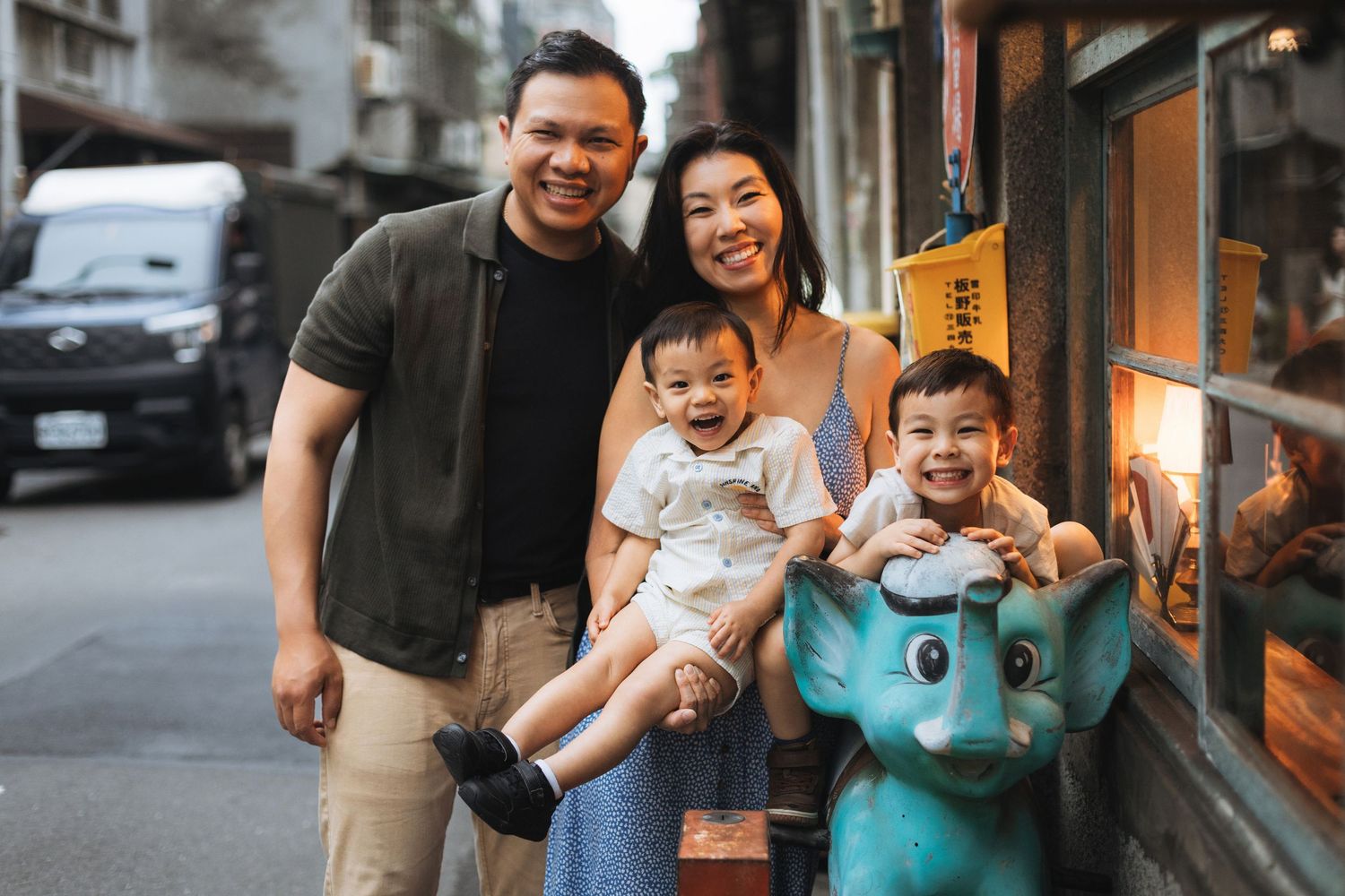 Happy Asian family of four posing on a charming street, parents smiling with two young children near a vintage elephant ride.
