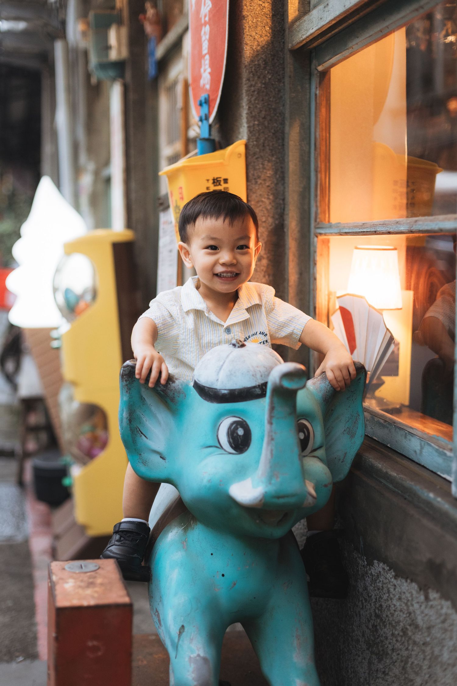 Smiling young Asian boy riding a turquoise elephant kiddie ride outside a shop with warm lighting.