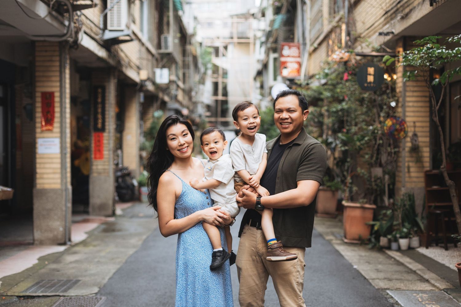 Happy Asian family of four posing on a charming narrow alley street, parents holding two young children, smiling outdoors.