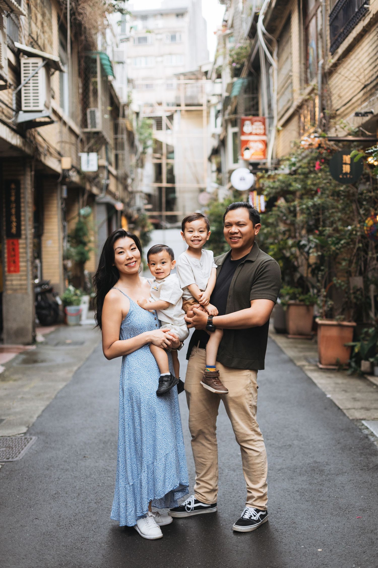 Happy Asian family of four posing together on a charming urban alleyway street, parents holding two young children.