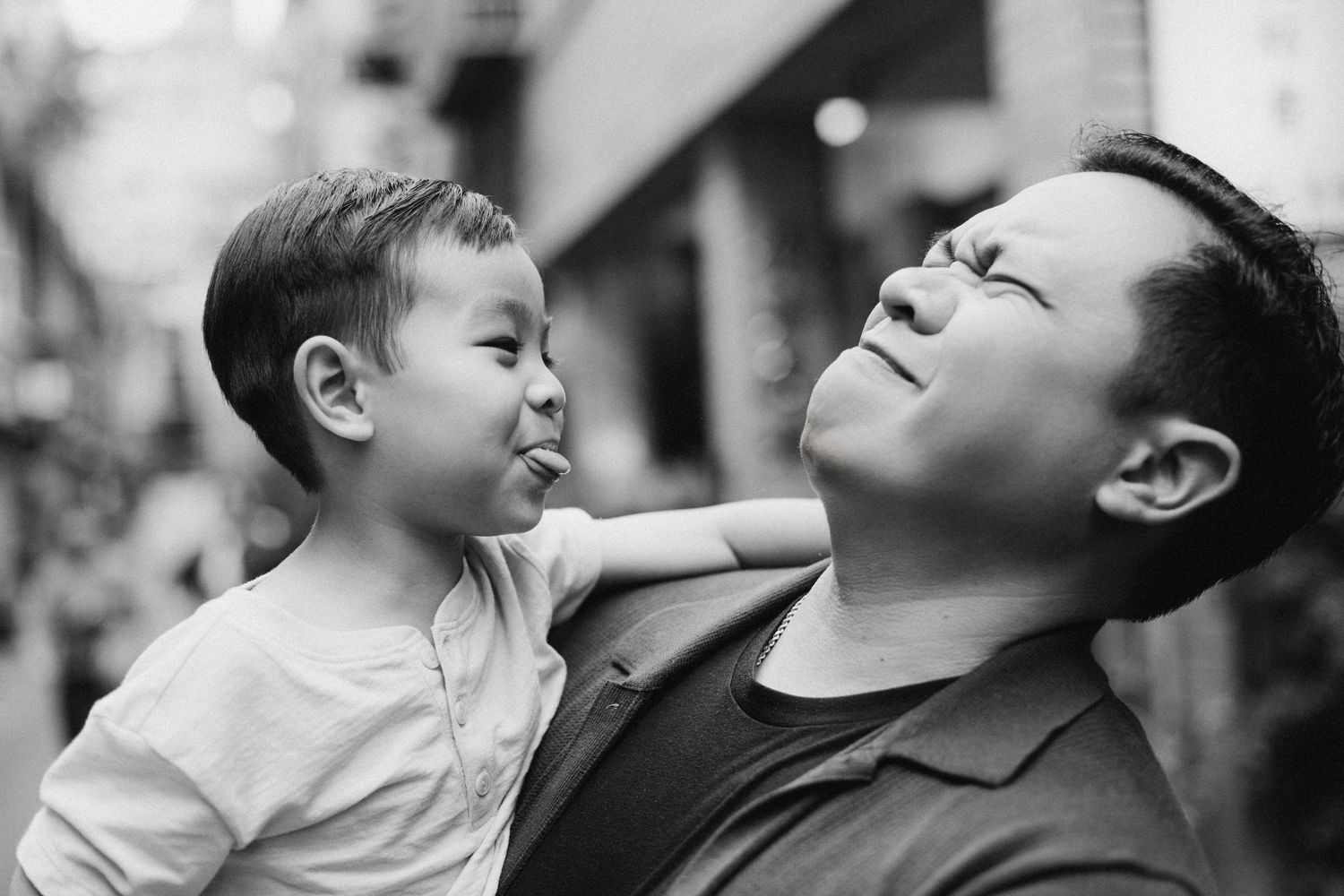 Black and white photo of a father holding his young son, who playfully sticks out his tongue while dad laughs.