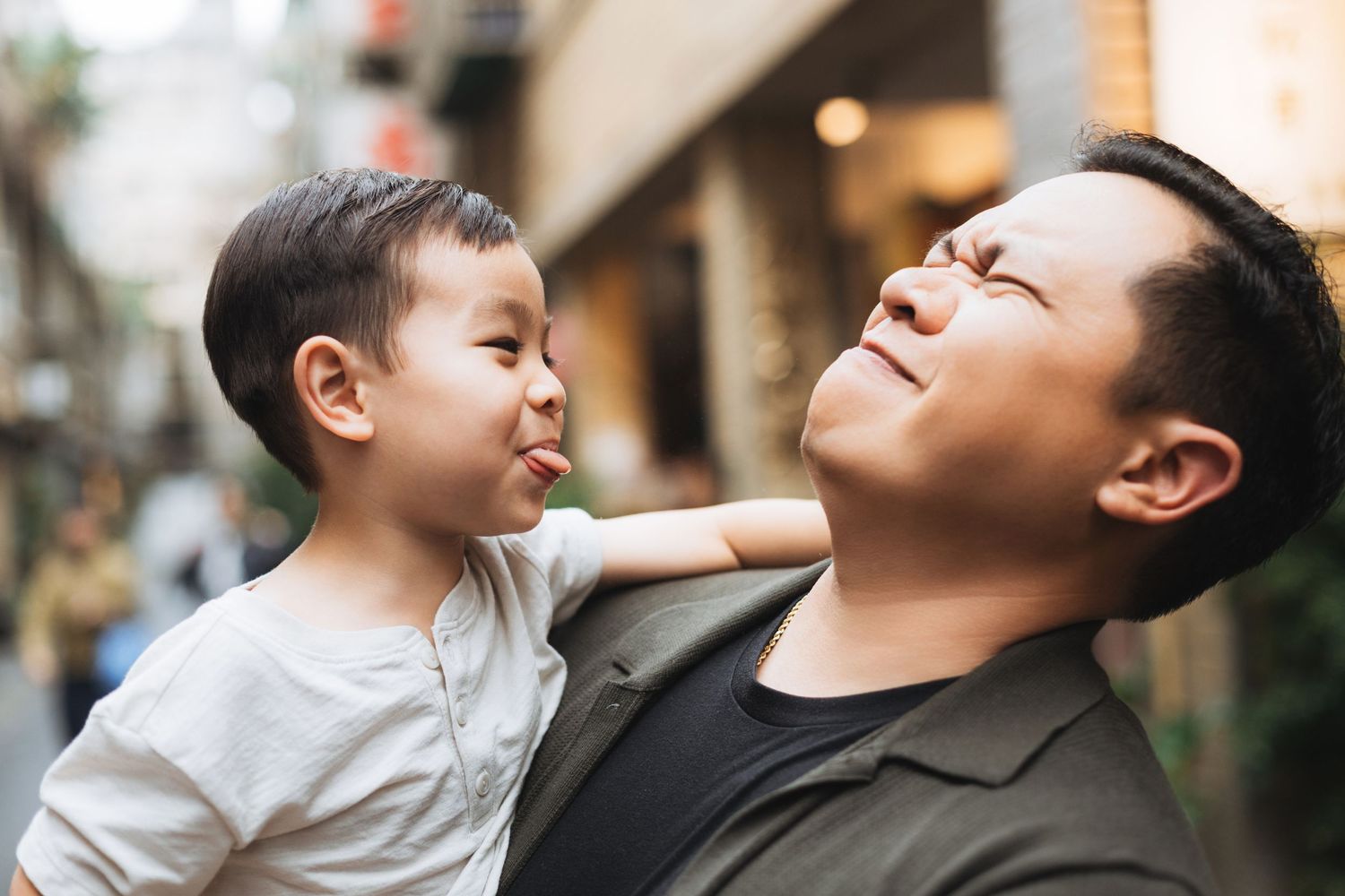 A father holds his young son who playfully sticks out his tongue while dad looks up smiling outdoors.