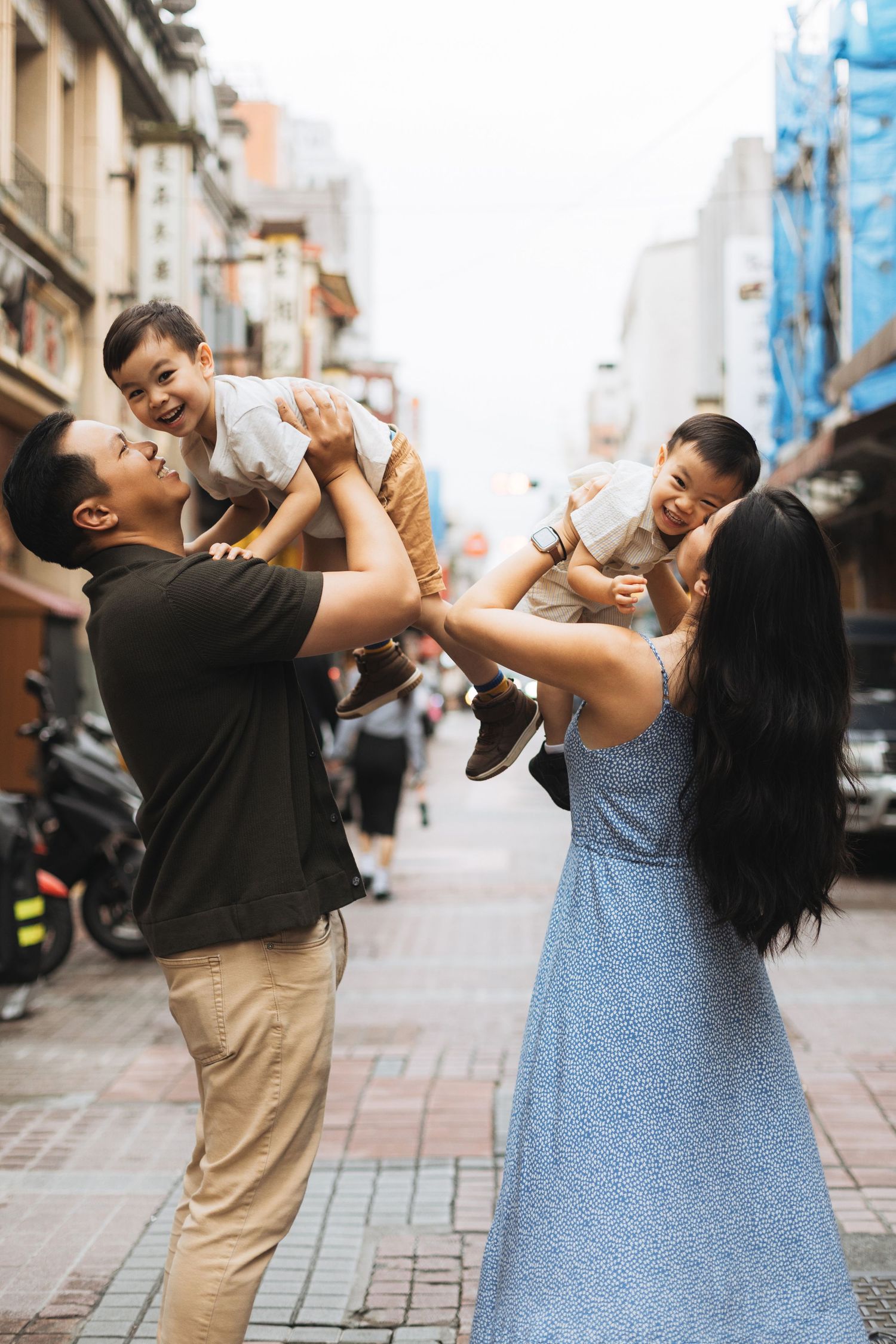 Happy Asian family lifting two young boys on a cobblestone street in an urban city setting.