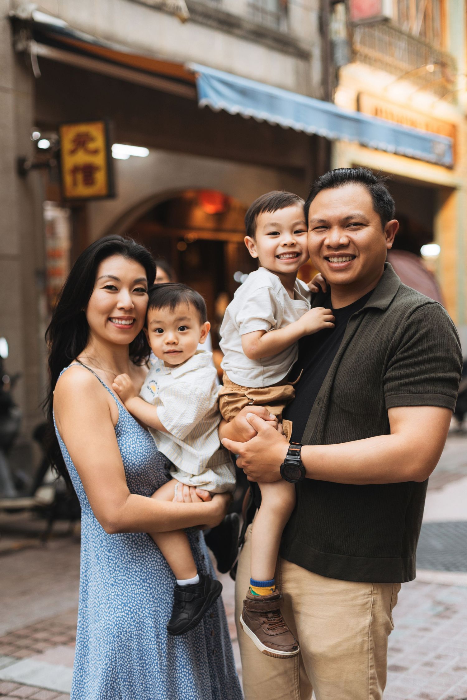 Happy Asian family of four posing on a city street, parents holding two young children, smiling outdoors.