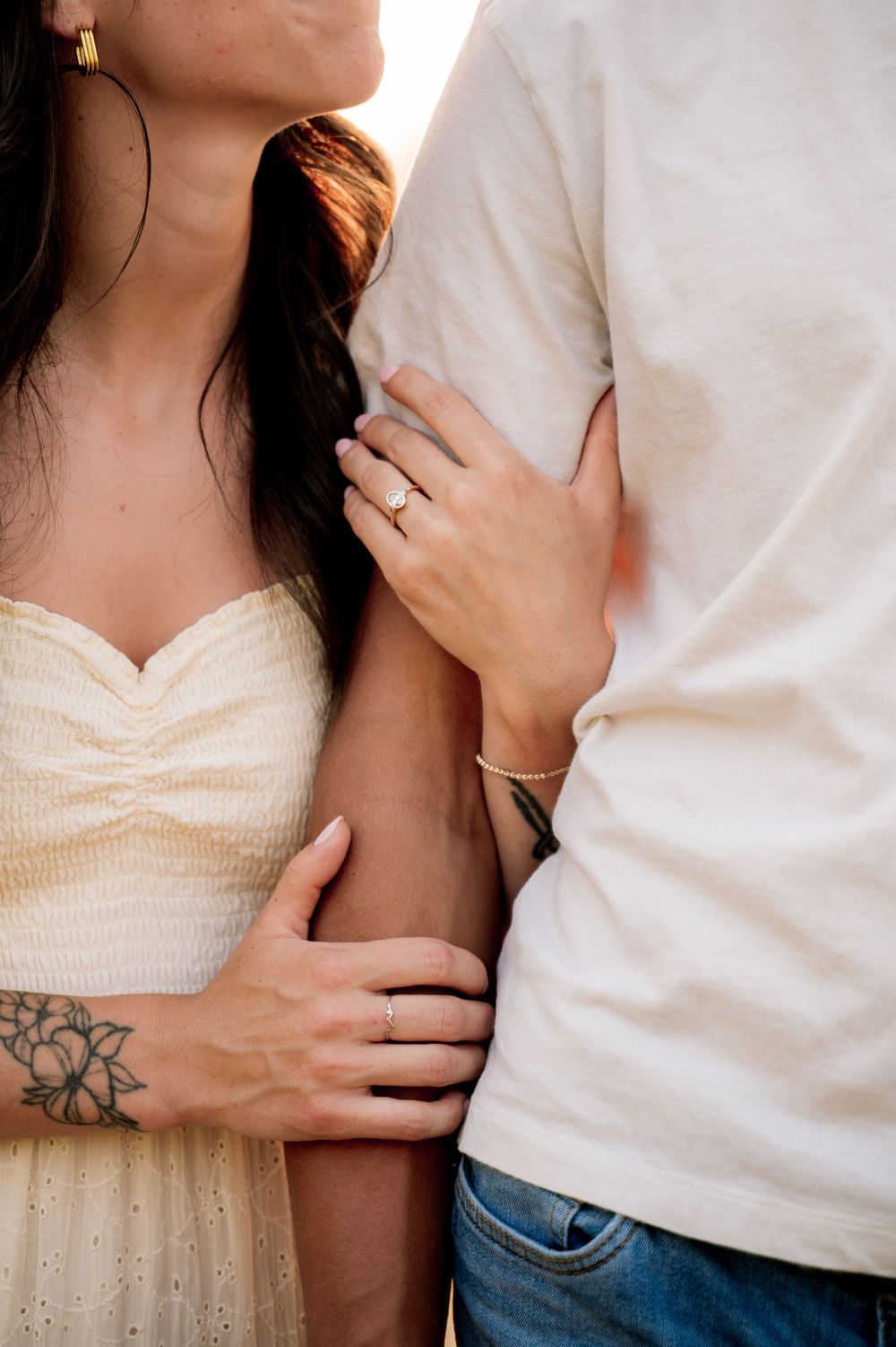 Close-up of an engaged couple embracing, showcasing a gold engagement ring and floral wrist tattoo.