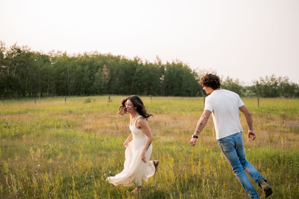 A joyful couple runs through a lush green meadow, woman in white dress, man in jeans, trees in background.