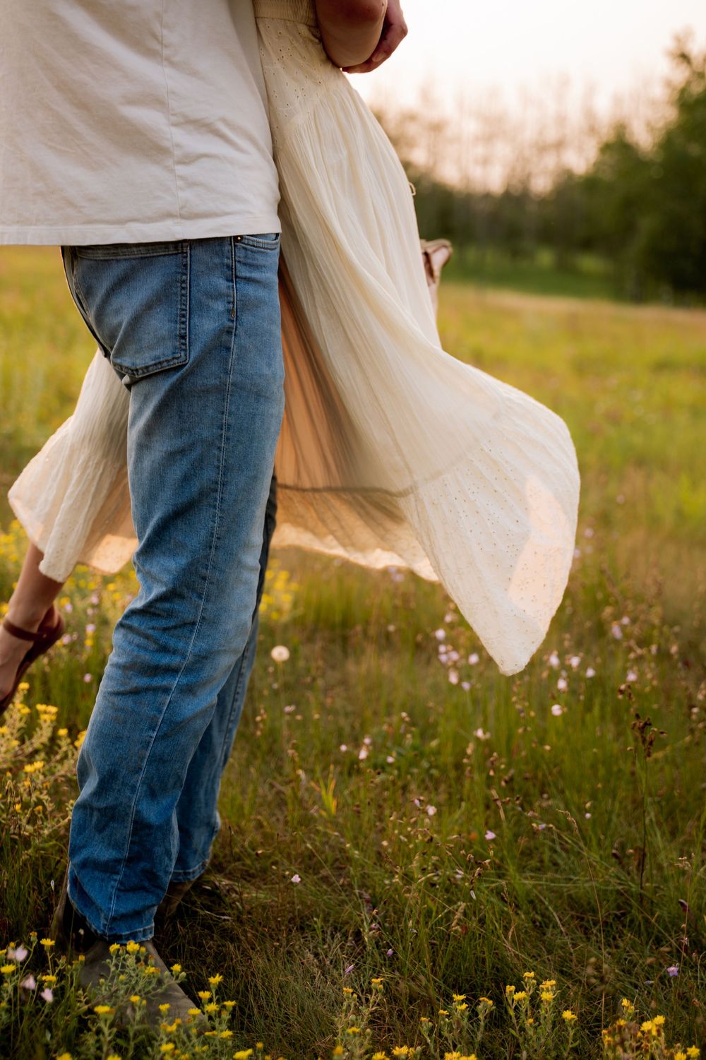 Couple walking through a sunny wildflower field, woman in flowing white dress twirling at sunset.
