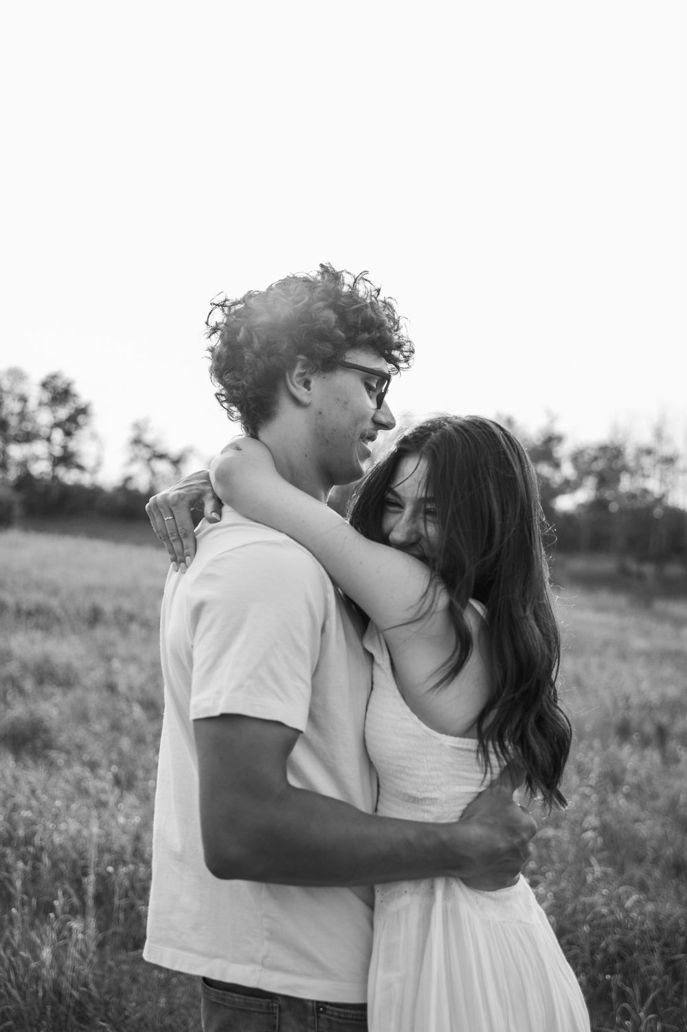 Black and white photo of a young couple embracing in a field, woman hugging man around neck outdoors.