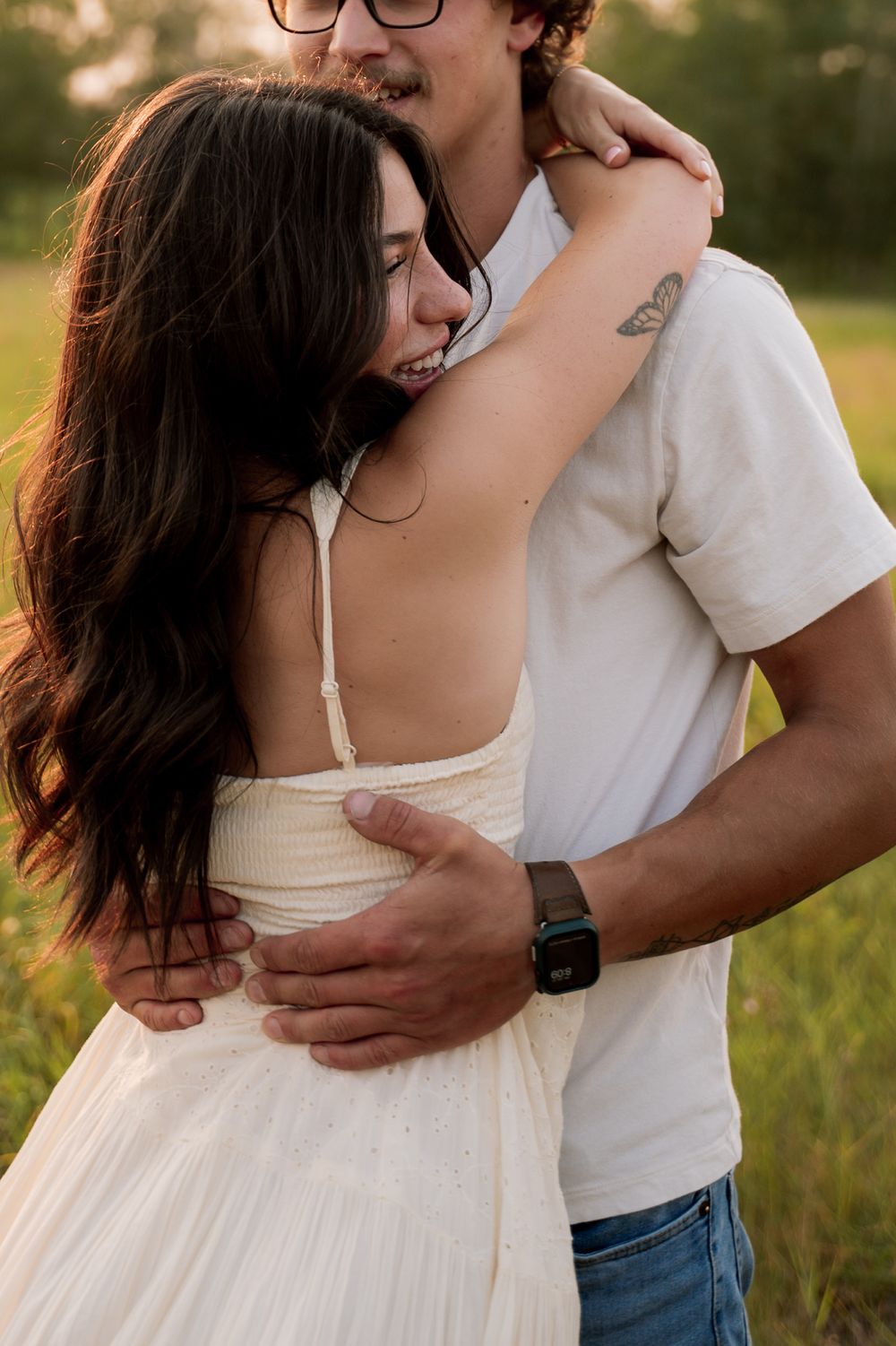 A couple embraces in a sunlit field, woman in white dress with long dark hair, man in white tee and jeans with a tattoo.