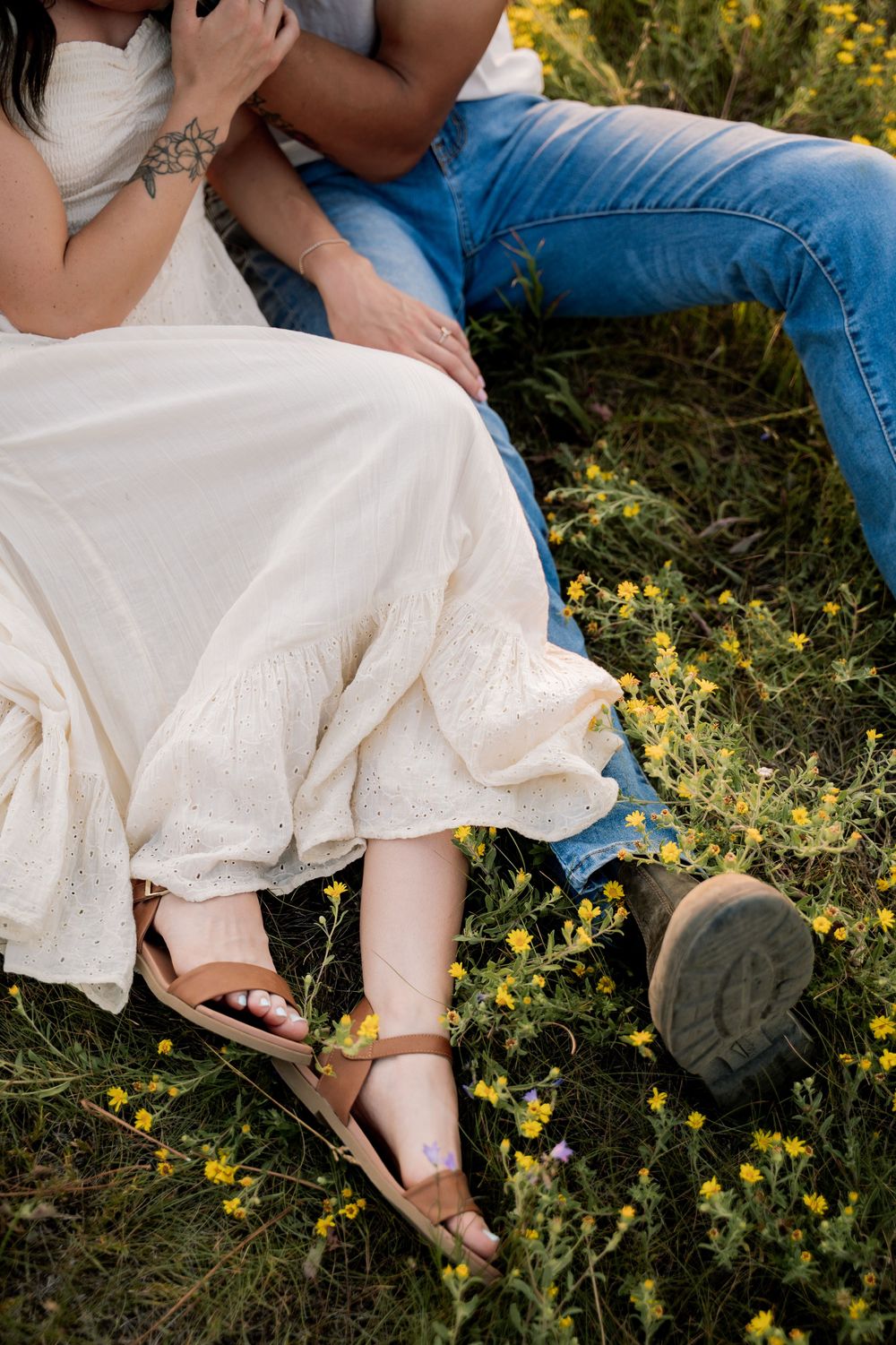 Couple sitting together in a wildflower meadow, woman in white dress and sandals, man in blue jeans.