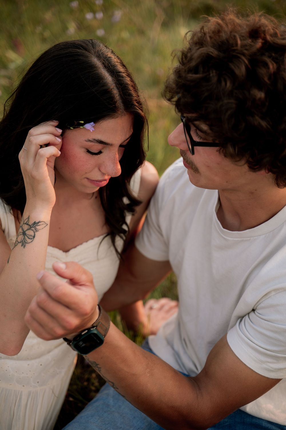 A young couple shares a tender moment outdoors, as a curly-haired man gazes at a dark-haired woman placing a flower in her hair.