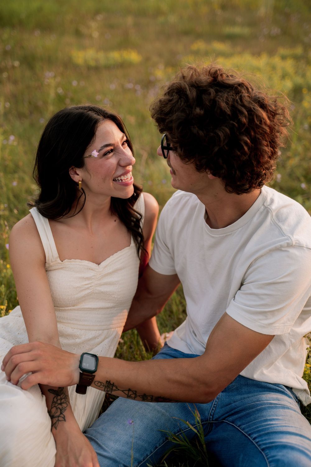Smiling couple sitting in a sunny wildflower field, woman in white dress laughing at curly-haired man.