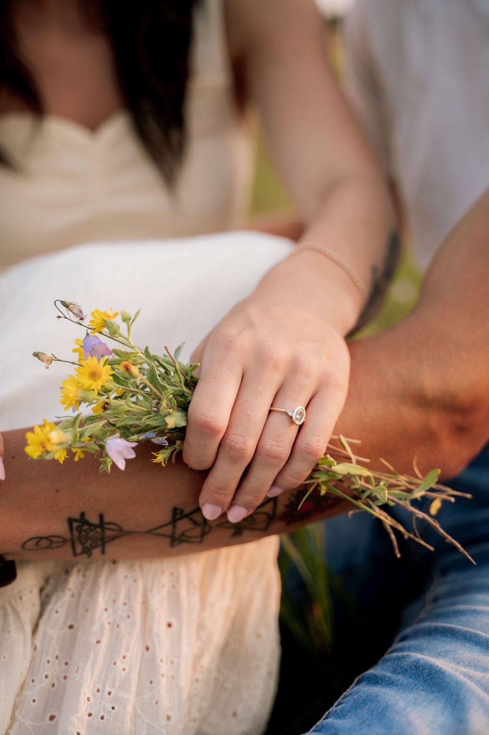 A woman's hand with an engagement ring rests on a tattooed arm holding wildflowers outdoors.
