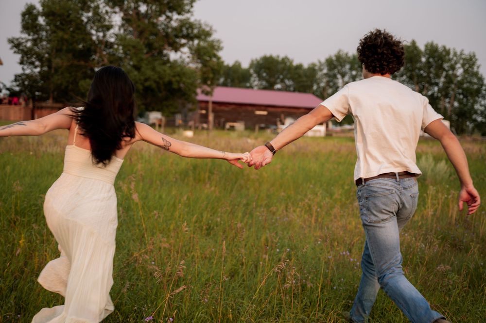 A bride in a white gown and groom in jeans hold hands running through a lush green field near a rustic barn.