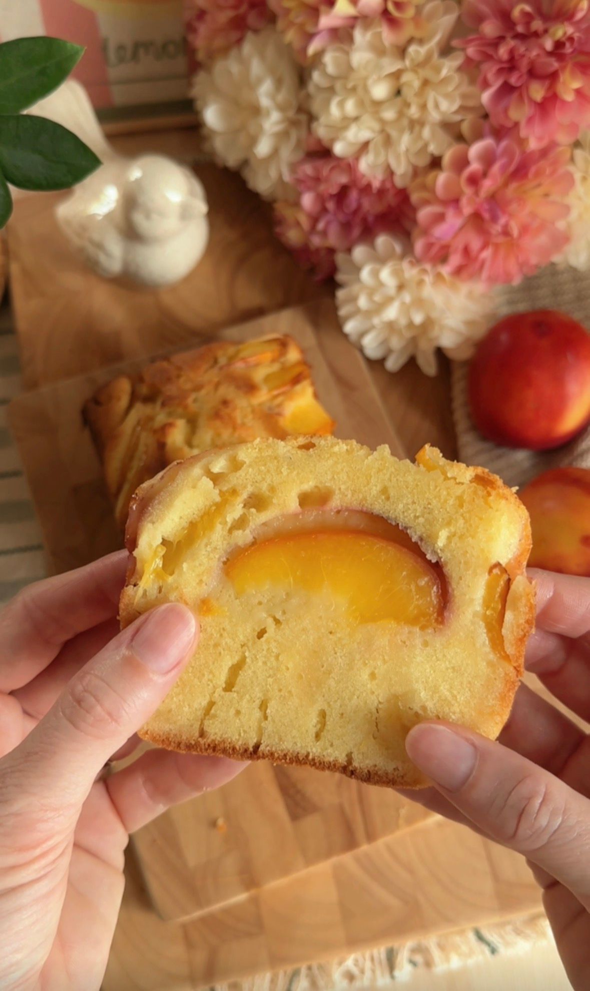 Golden peach loaf cake in parchment-lined pan, fresh peaches and pink flowers on wooden board.