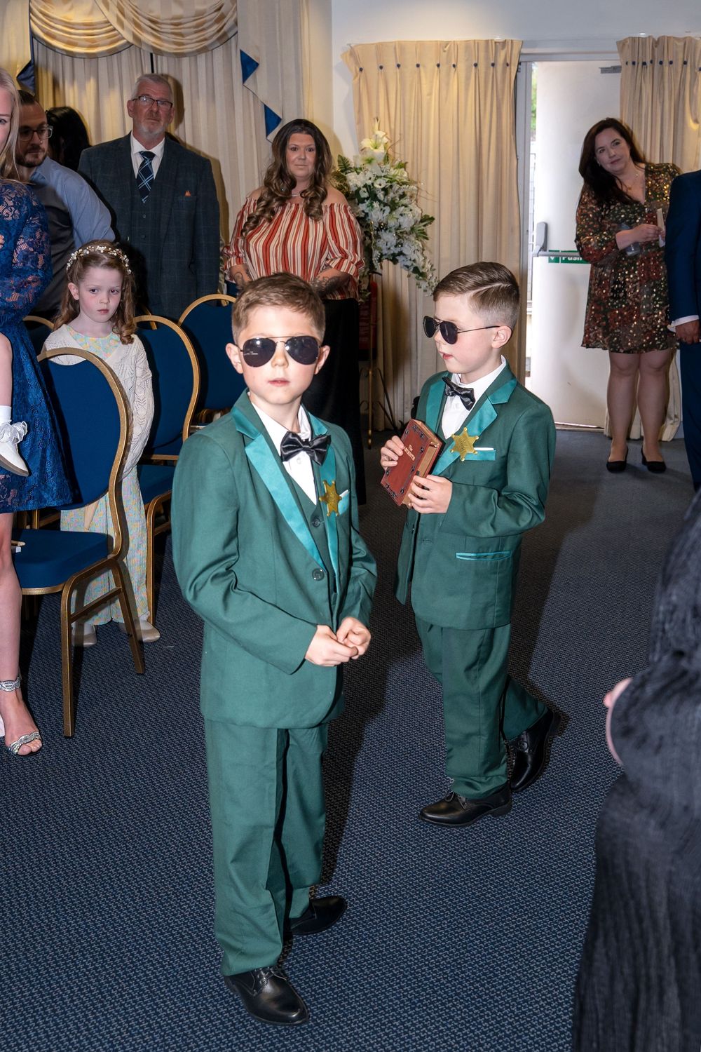 Two young boys in matching green suits and sunglasses walk down the aisle at a wedding ceremony.
