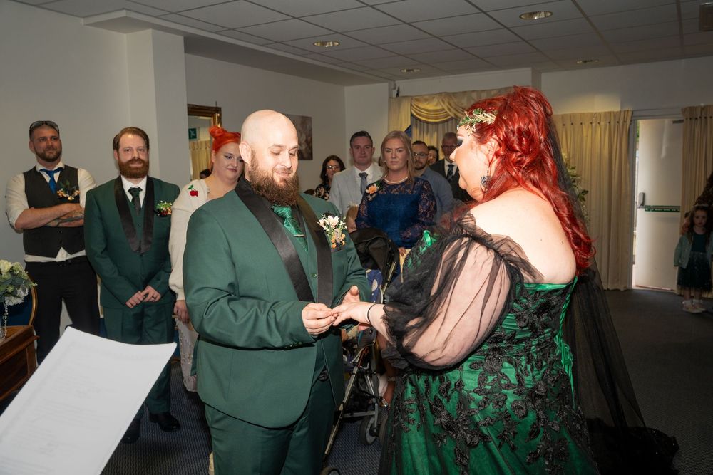 Couple in matching green outfits exchange rings during indoor wedding ceremony with guests watching.