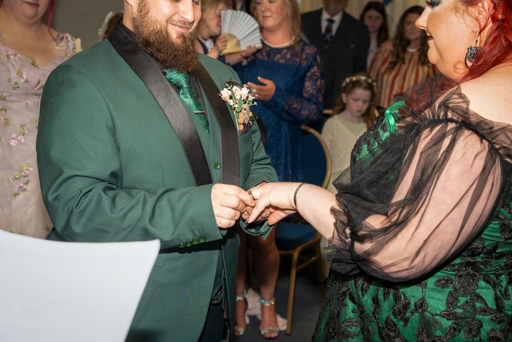 Groom in green suit exchanging rings with bride in emerald dress during wedding ceremony.
